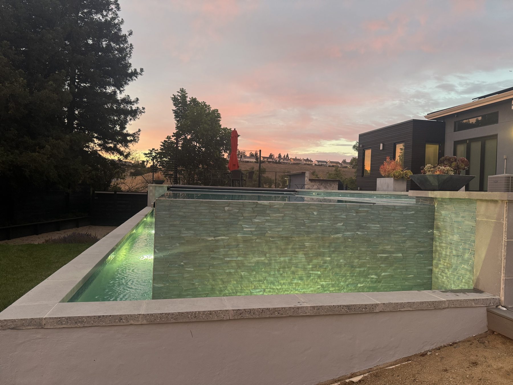 Raised vanishing-edge pool with its glass-tile wall and catch basin glowing under a pink sunset